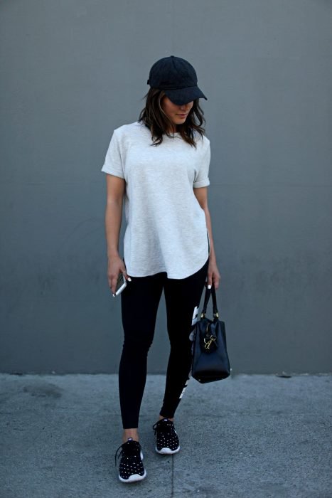 Chica en pared gris, con blusa blanca holgada, leggins negro, tenis deportivos, cachucha negra y en mano su celular y bolso negro