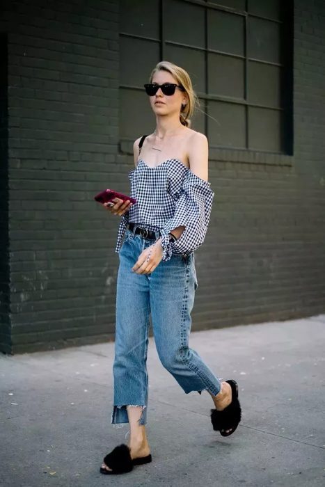 Chica caminando por la calle con el celular en la mano mientras usa jeans, campesina y unas sandalias peludas 