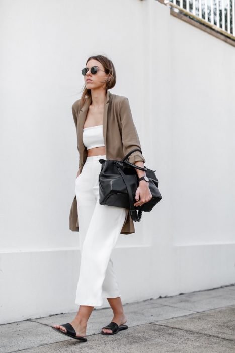Chica usando un outfit blanco con un cardigan de color café, bolso negro y sandalias de color negro mientras camina por la calle 