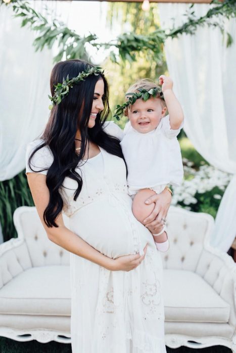 Vestidos para baby shower, mujer embarazada cargando a su bebé con ropa que combina, mamá con vestido blanco y corona de flores y bebé con ropón blanco
