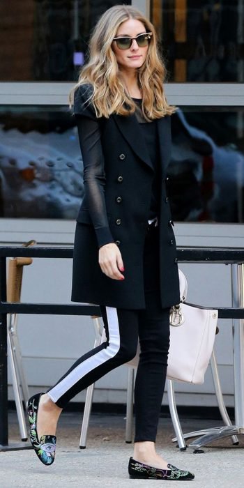 Chica caminando por la calle, con leggins negros con detalle blanco a los lados, blusa negra y zaco negro, con bolsa beige en mano