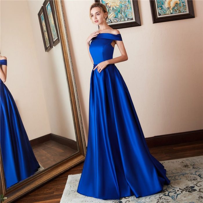 Chica rubia frente al espejo con vestido de fiesta largo color azul royal satinado sin hombros