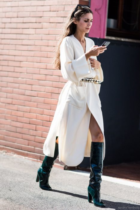 Chica usando un vestido blanco y botas de charol verde mientras camina por la calle mirando su celular