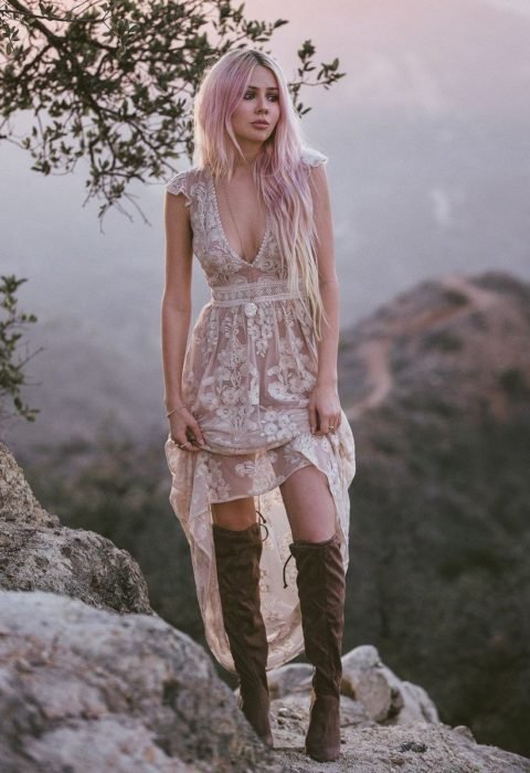 Ropa estilo boho o hippie chic; mujer en la montaña, de cabello largo y rosa, con vestido de encaje y botas largas cafés