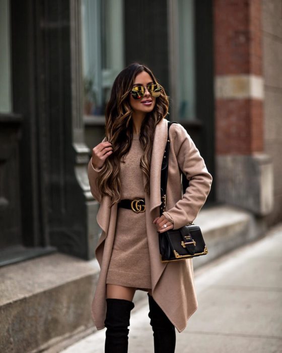 Chica usando un vestido y abrigo en color café con un cinturón chanel y botas mientras camina por la calle 