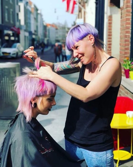 una mujer con corte 'bowl' en morado le corta el cabello a otra mujer con pelo rosa en una banqueta