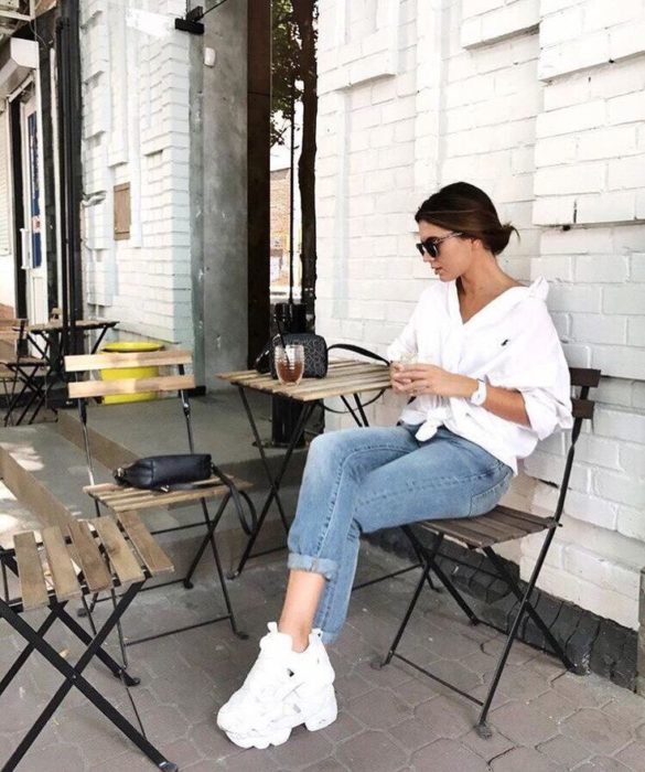 Chica sentada en un café mientras usa unos pantalones de mezclilla, blusa blanca y tenis fila de color blanco 