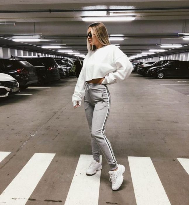 Chica parada en medio de un estacionamiento usando un pantalón de color girs, suéter blanco y tenis fila blancos 