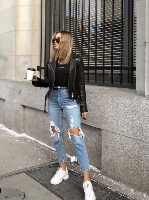Chica parada en medio de la calle sosteniendo un café mientras usa jeans desgarrados, chaqueta y blusa negra y tenis fila de color blanco 