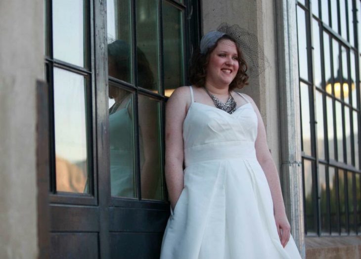 Una novia recargada en una puerta de un edificio posa con una mano dentro del bolsillo de su vestido y la otra a un costado de su cuerpo