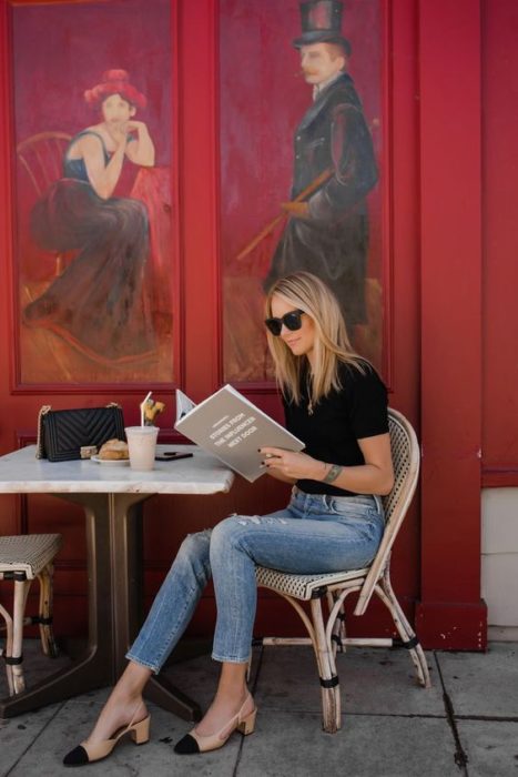 Mujer rubia sentada leyando la carta de un restaurante viste blusa negra, jeans, lentes de sol y zapatos cuadrados