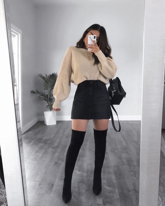 Chica se toma selfie frente al espejo con suéter beige, falda negra circular y botas altas negras