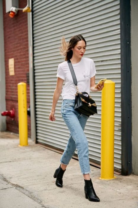 Chica camina por la calle vistiendo jeans, blusa blanca y botines negros